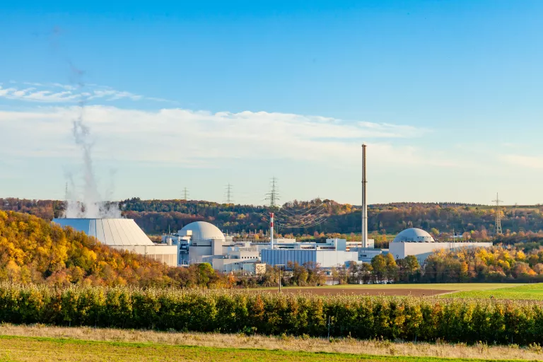 Blick auf einen Gebäudekomplex unter blauem Himmel und leichter Bewölkung. Am linken Bildrand ist ein Kühlturm zu erkennen, aus dem Dampf aufsteigt. Im Bildvordergrund zeichnen sich ein schmales Waldstück, Wiesen und Felder sowie ein bewaldeter Hügel ab. Im Bildhintergrund sind weitere flache bewaldete Hügel sowie Strommasten zu erkennen.