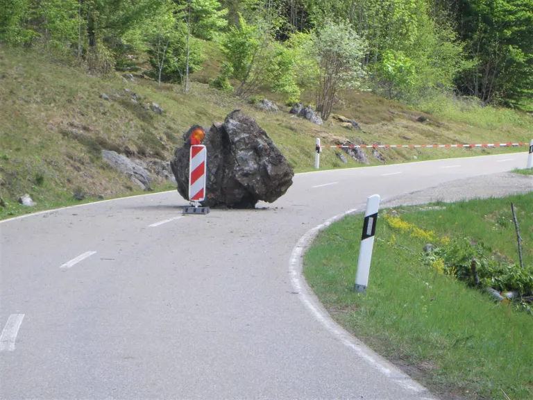 Ansicht eines PKW-großen Blocks, der mittig auf einer Landesstraße liegt. Vor dem Block steht eine eine Warnbarke.