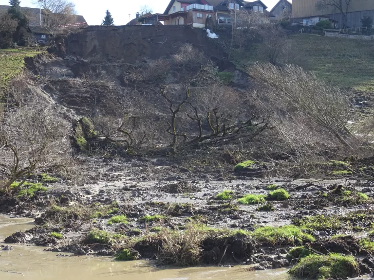 Ansicht einer ausgedehnten Rutschung. Im Vordergrund ist das durchnässte, breiig, schlammige Rutschmaterial zu sehen. Darin befinden sich einige umgestürzte Bäume und zahlreiche Grasfetzen. Im Hintergrund sind die steile Abrisskante der Rutschung und ein unmittelbar daran anschließendes Wohngebäude sichtbar.