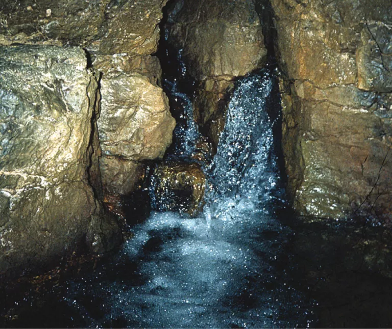 Aus einer Karstspalte zwischen eng stehendem, dunklem Festgestein tritt Quellwasser hervor