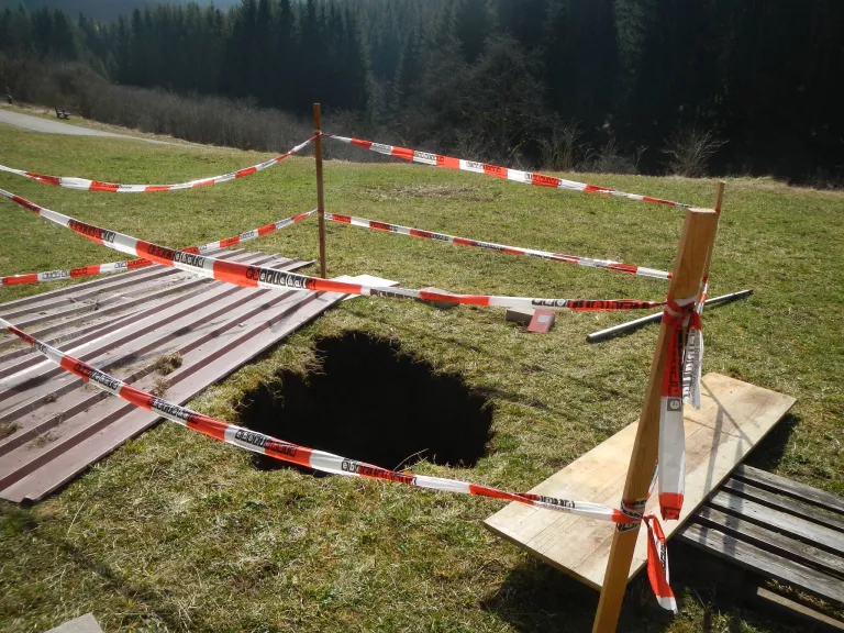 Blick auf ein flaches grasbewachsenes Hanggelände im Bildvordergrund und dunkler Nadelwald im Bildhintergrund. Etwa mittig in der unteren Bildhälfte ist ein ca. 1 m großes schwarzes Loch im Boden zu erkennen. Dies ist mit an vier Holzlatten befestigtem Flatterband vor Betreten gesichert. Rechs und links des Lochs liegen eine Schaltafel sowie zwei Dachbleche zum Abdecken des Lochs auf dem Boden. Diese sind nun zur Seite geräumt. 