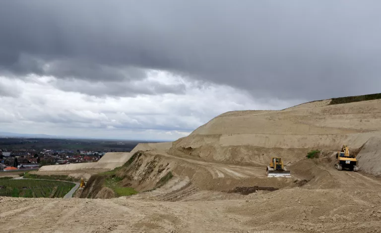 Im hellen Löss wurden große Terrassen aus der Landschaft herausmodelliert. Drei Baustellenfahrzeuge sind zu sehen. Am Unterhang sind Weinberge zu erkennen und dahinter eine Siedlung. Im fernen Hintergrund sieht man am Horizont ein Mittelgebirge. Der Himmel ist mit dunklen Wolken verhangen.