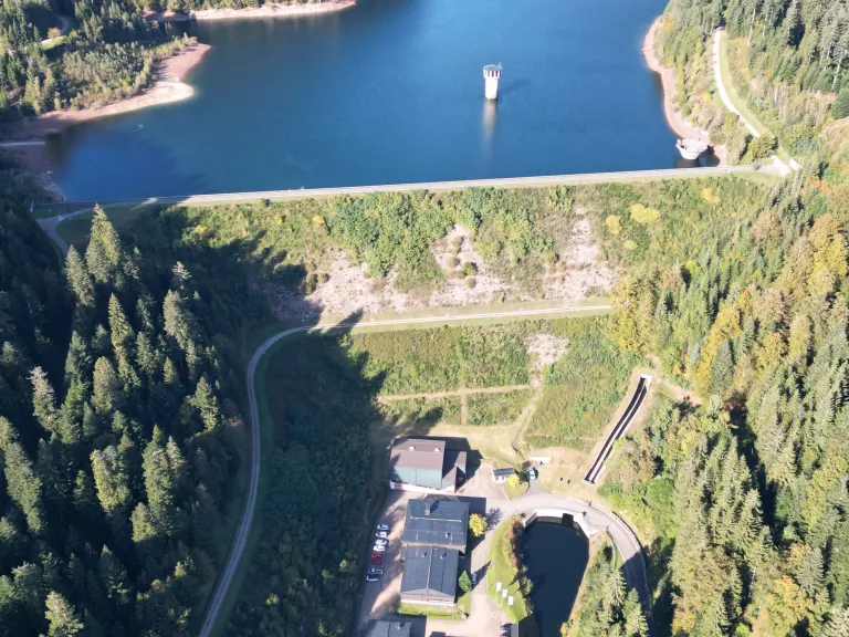 Blick auf einen teils mit Gras bewachsenen Dammkörper vor einem Stausee. In der Wasserfläche sind technische Anlagen der Trinkwassertalsperre zu erkennen. Im Bildvordergrund unterhalb der Staumauer zeichnen sich Gebäude der Talsperre ab.