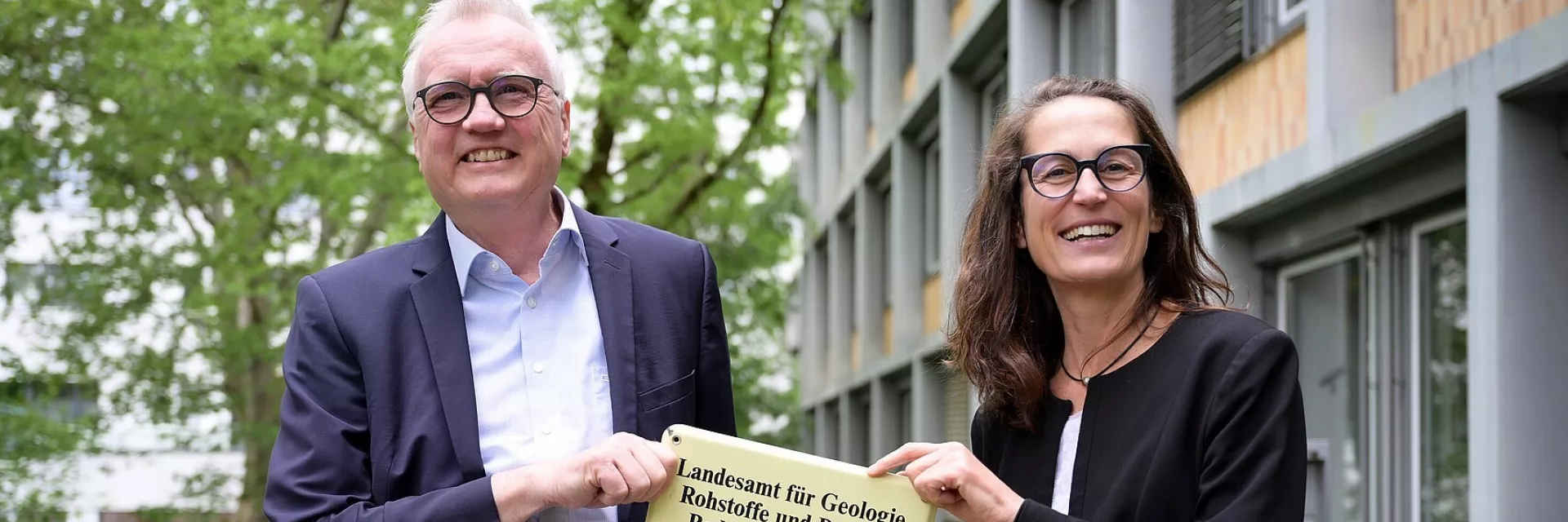 links ein weißhaariger, lächelnder Herr mit Brille - rechts eine dunkelhaarige, lächelnde Frau mit Brille; beide halten gemeinsam ein Schild des LGRB