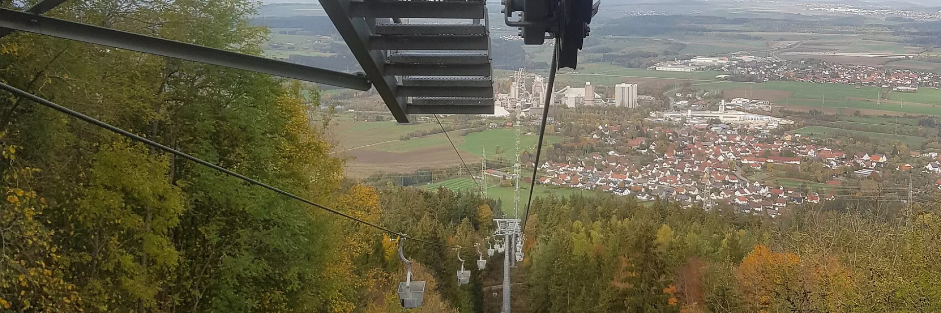 Blick entlang einer Seilbahn nach unten durchLaubwald, im Hintergrund eine Ortschaft und eine Industrieanlage