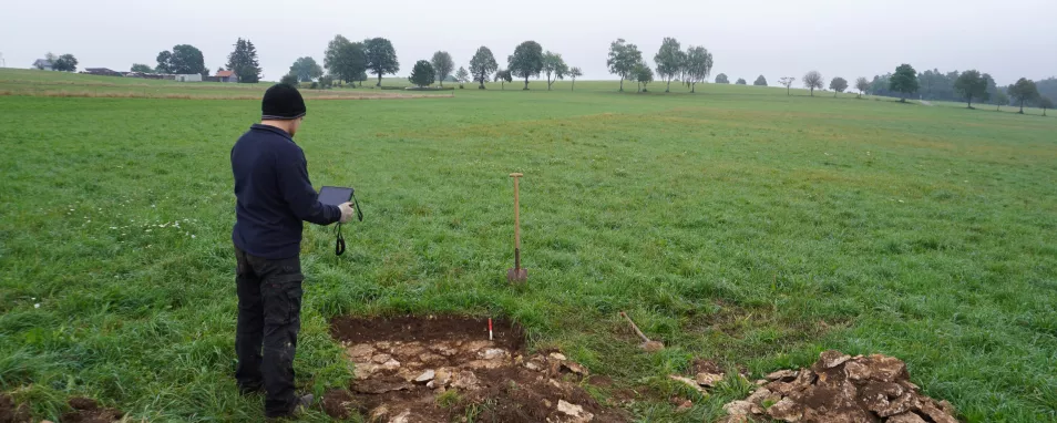 Auf einer Wiese ist ein flaches Bodenprofil aufgegraben. Direkt unter humosem Oberboden folgt heller Kalkstein. Dabei steht ein Mann, der mit einem Gelände-Tablet Bodendaten erfasst. Im Hintergrund stehen einzelne Bäume. 